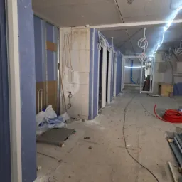 Wnętrze w trakcie prac wykończeniowych: widoczne profile metalowe, płyty karton-gips w kolorze niebieskim, wystające przewody elektryczne i rury, surowy beton na podłodze i suficie, oraz zwisające...