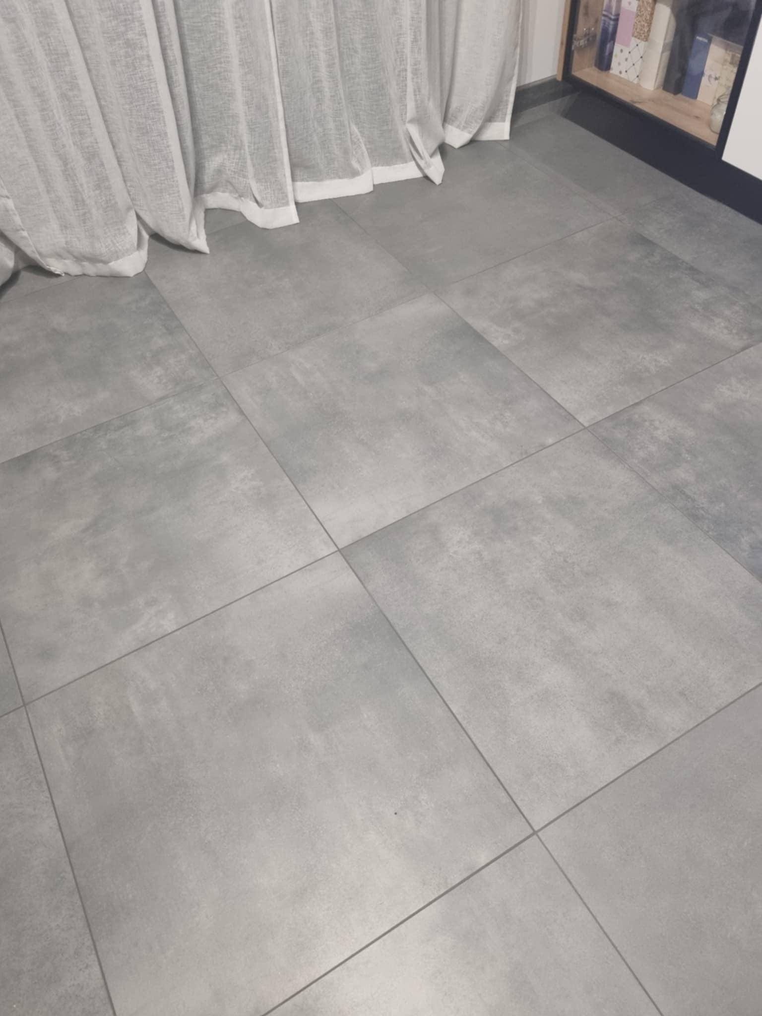 Szare, matowe płytki podłogowe ułożone w kwadraty, widoczne fragmenty białych zasłon i półki z kosmetykami.