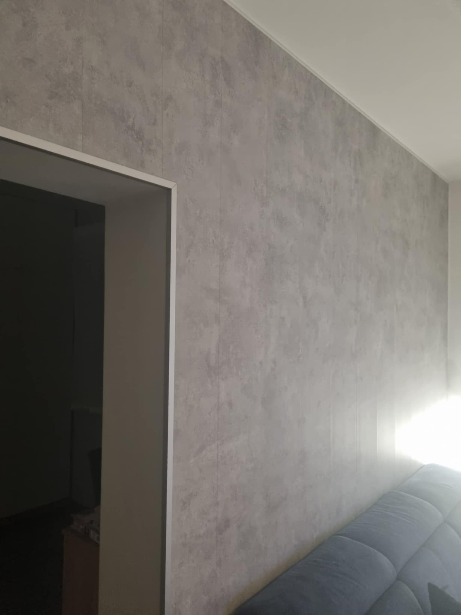 Ściana wykończona szarą, imitującą beton gładzią, z widocznym wejściem do pomieszczenia po lewej stronie i fragmentem sofy w dolnym rogu.