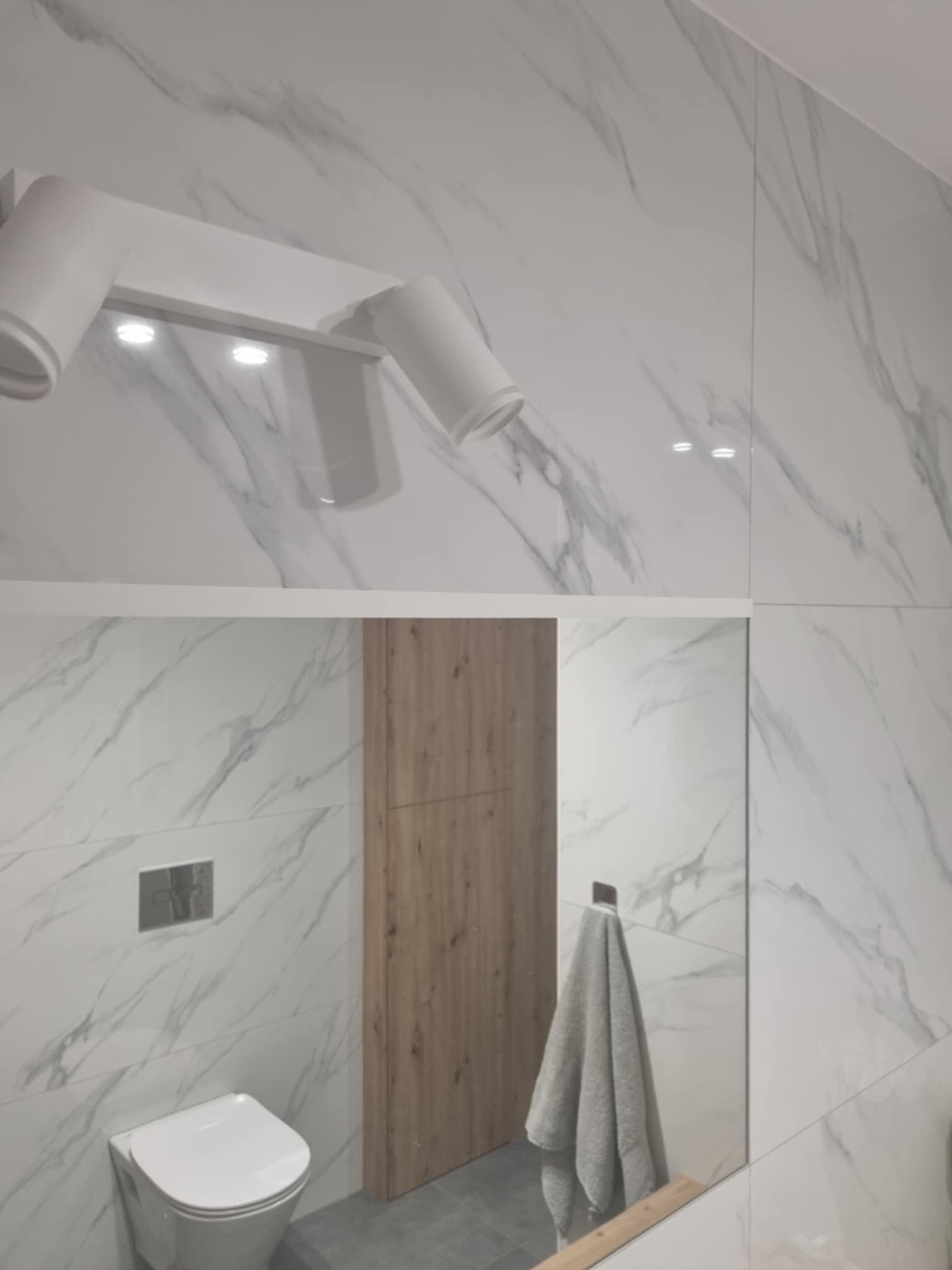 Nowoczesna łazienka z białymi płytkami imitującymi marmur, drewnianymi drzwiami, wiszącą toaletą i szarą wiszącą szmatką, oświetlona trzema reflektorami.