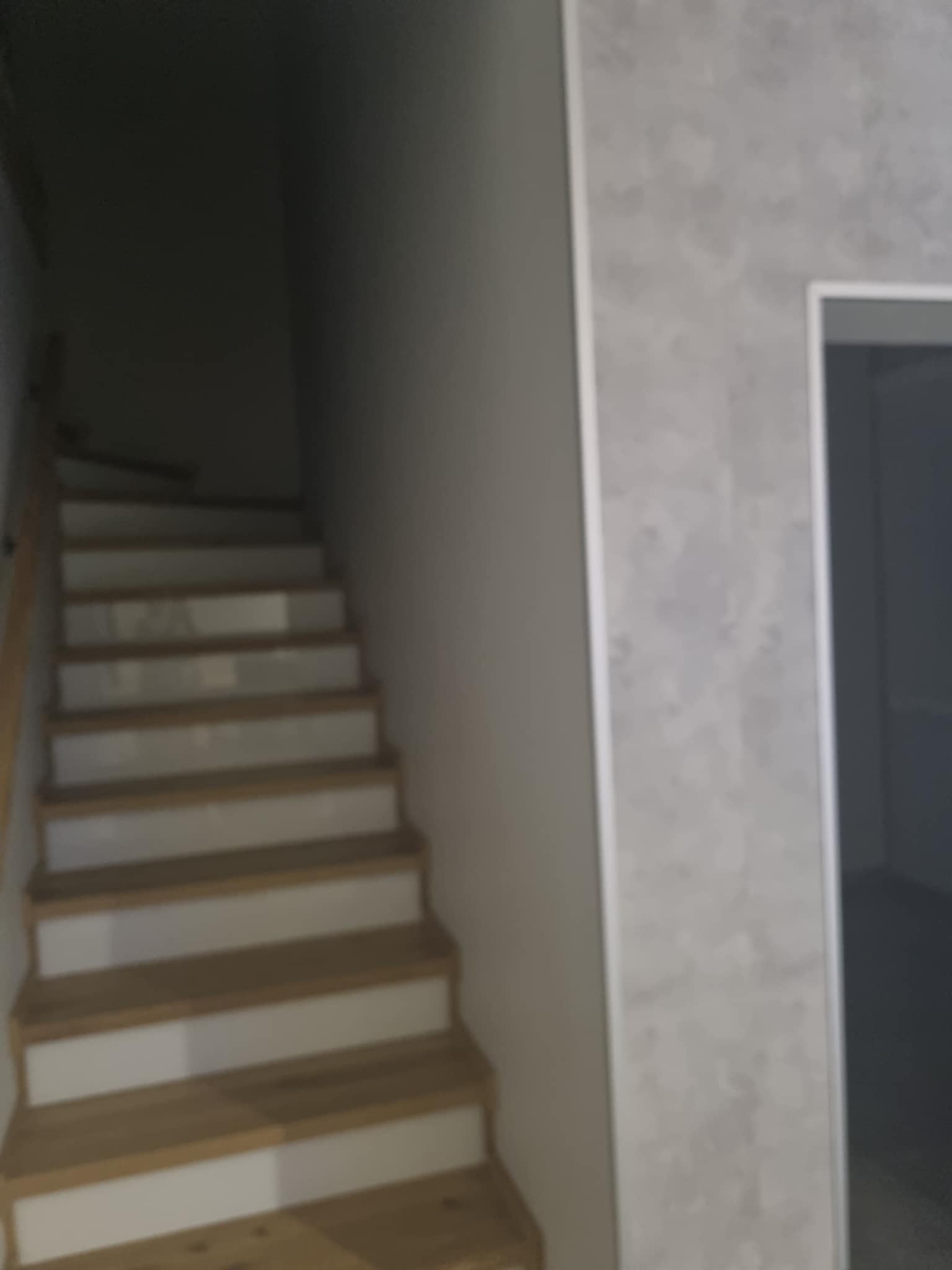 Schody drewniane z białymi podstopnicami w słabo oświetlonym wnętrzu, widoczna ściana z dekoracyjną strukturą i fragment ościeżnicy drzwi.