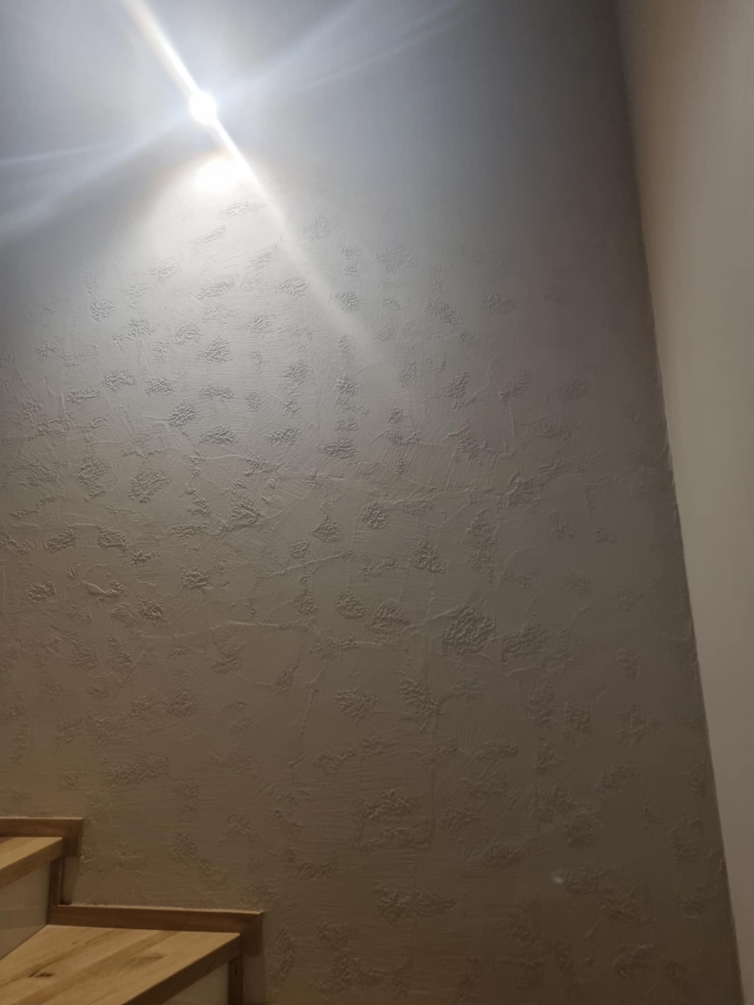 Szara ściana z dekoracyjną fakturą przy schodach, oświetlona górnym światłem, tworząca efekt cienia i światła na powierzchni.