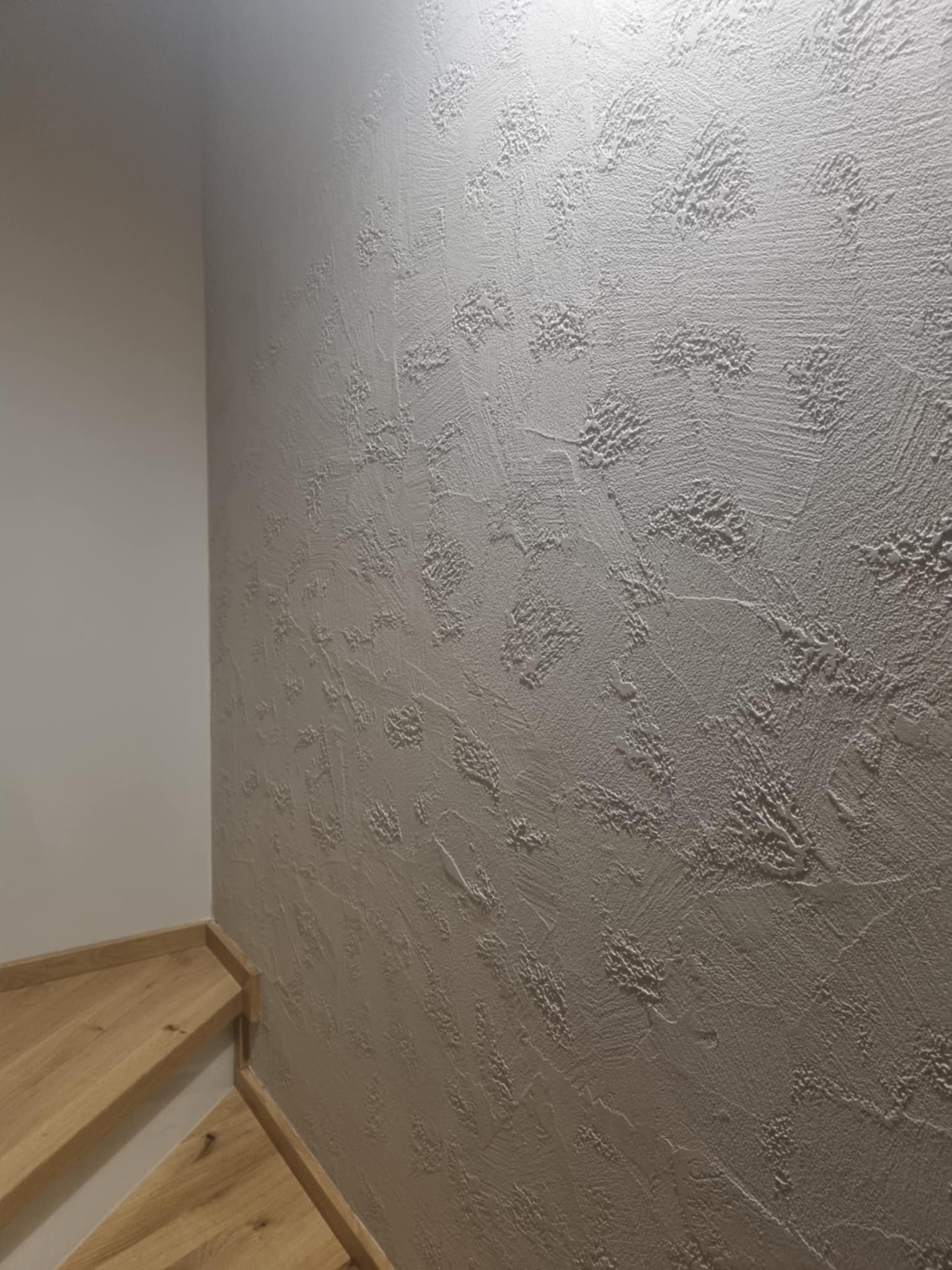 Ściana pokryta dekoracyjną, strukturalną gładzią w odcieniach szarości, przylegająca do drewnianych schodów.