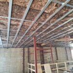 Sufit podwieszany w trakcie montażu: widoczna metalowa konstrukcja szkieletowa, wypełniona wełną mineralną. W tle częściowo widoczne regały i ściana z surowego betonu.