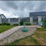 Nowoczesny dom jednorodzinny z szarą elewacją, widok z ogrodu z kamienną ścieżką i ozdobnymi kamieniami. Metalowe ogrodzenie panelowe z betonowymi słupkami. Pochmurne niebo.