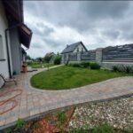 Ułożona kostka brukowa tworząca ścieżkę wzdłuż domu, z widokiem na trawnik, ogrodzenie i dom w tle, pochmurne niebo.