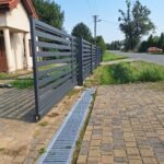 Nowoczesne, poziome, metalowe ogrodzenie posesji w kolorze antracytowym, z widocznym odwodnieniem liniowym i brukowaną ścieżką, na tle zieleni i domu jednorodzinnego.