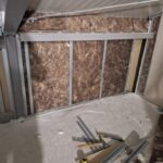 Ściana z metalowym stelażem wypełniona izolacją z wełny mineralnej, widoczne elementy konstrukcyjne i fragmenty płyty gipsowo-kartonowej, surowy stan wykończenia.