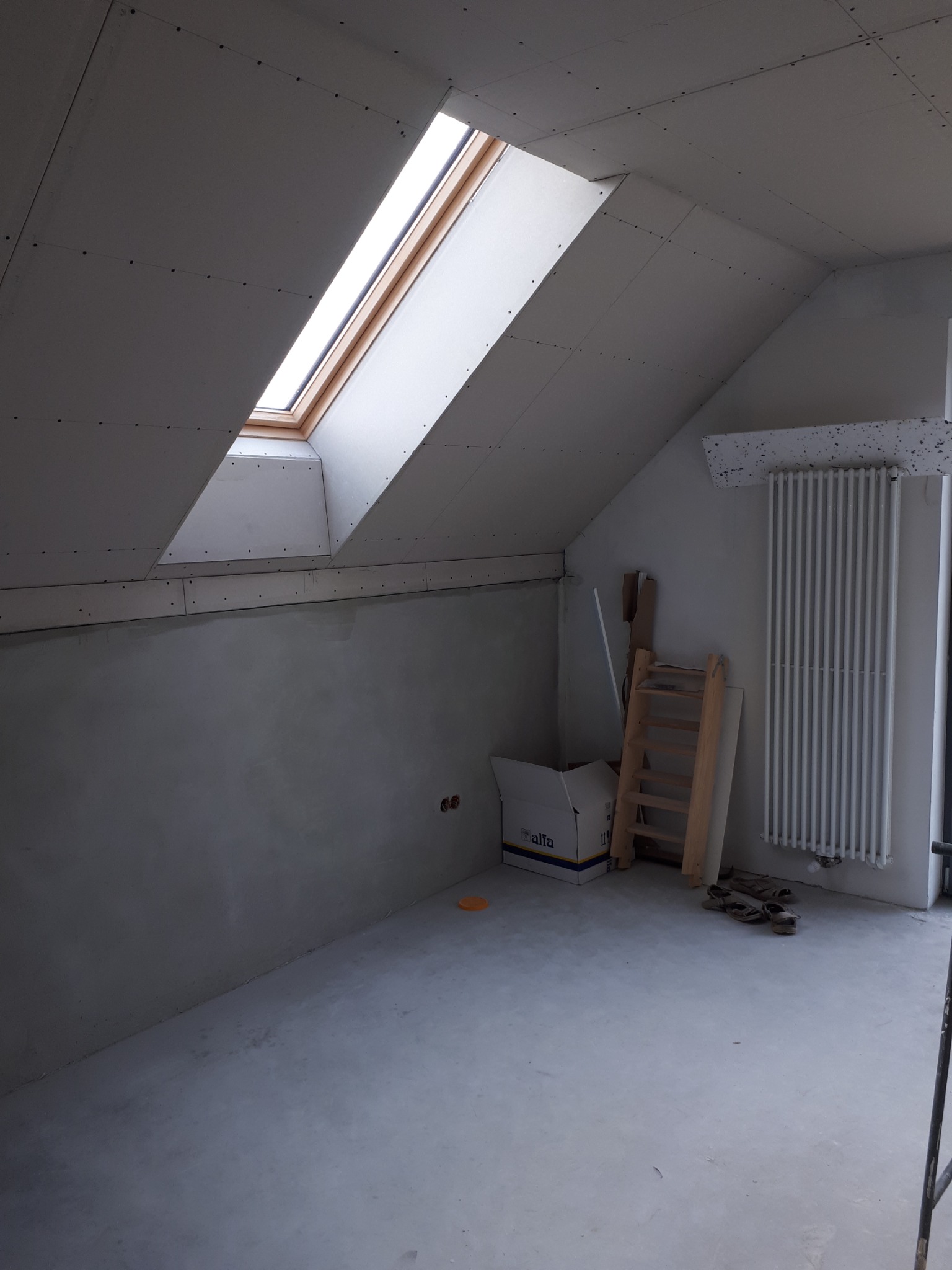 Poddasze w trakcie prac wykończeniowych: zamontowana płyta karton-gips na skosach, okno dachowe z drewnianą ramą, ściany przygotowane pod gładzie, grzejnik żeberkowy i rozłożone robocze obuwie.