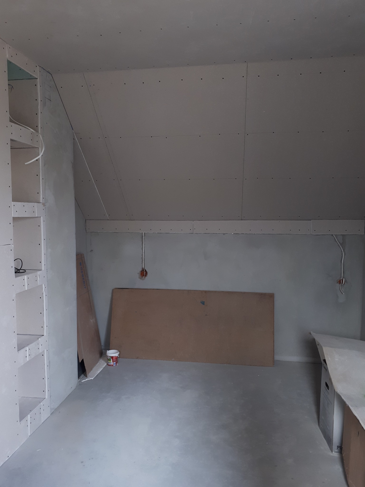 Poddasze w trakcie adaptacji: ściany i sufit pokryte płytami kartonowo-gipsowymi, widoczne kable elektryczne, konstrukcja regału z płyty GK, płyta MDF oparta o ścianę, małe wiaderko z farbą przy...
