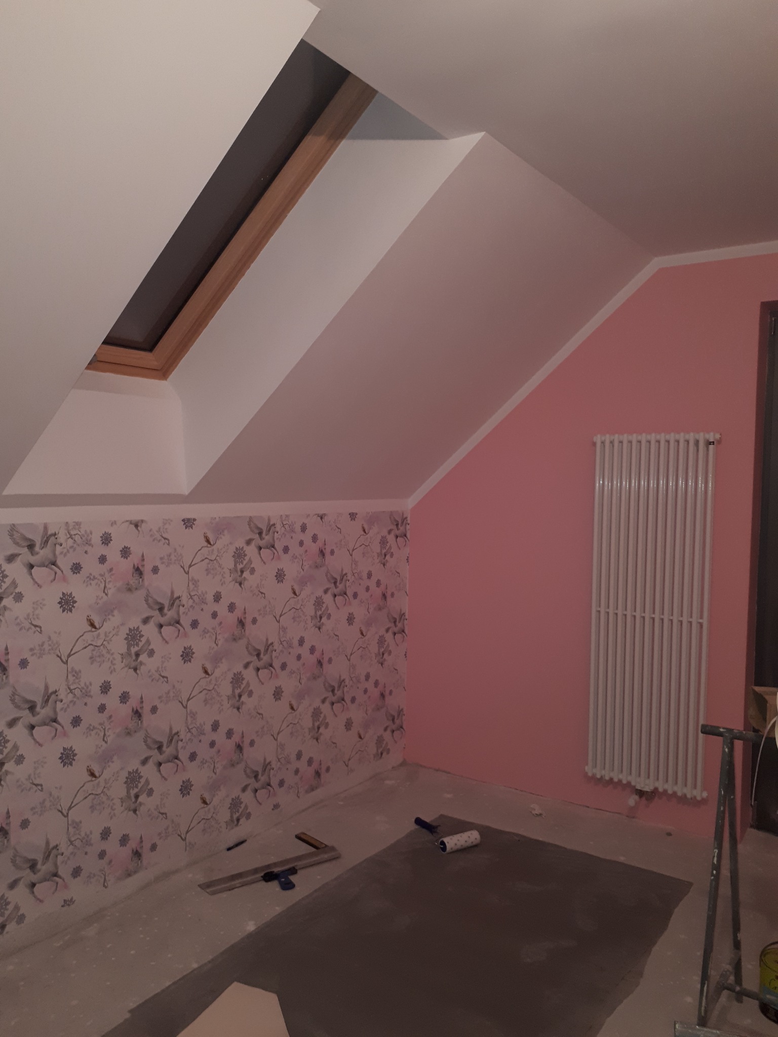 Pokój na poddaszu w trakcie odświeżania: ściana z tapetą w jednorożce, różowa ściana z grzejnikiem, narzędzia malarskie na podłodze zabezpieczonej folią.