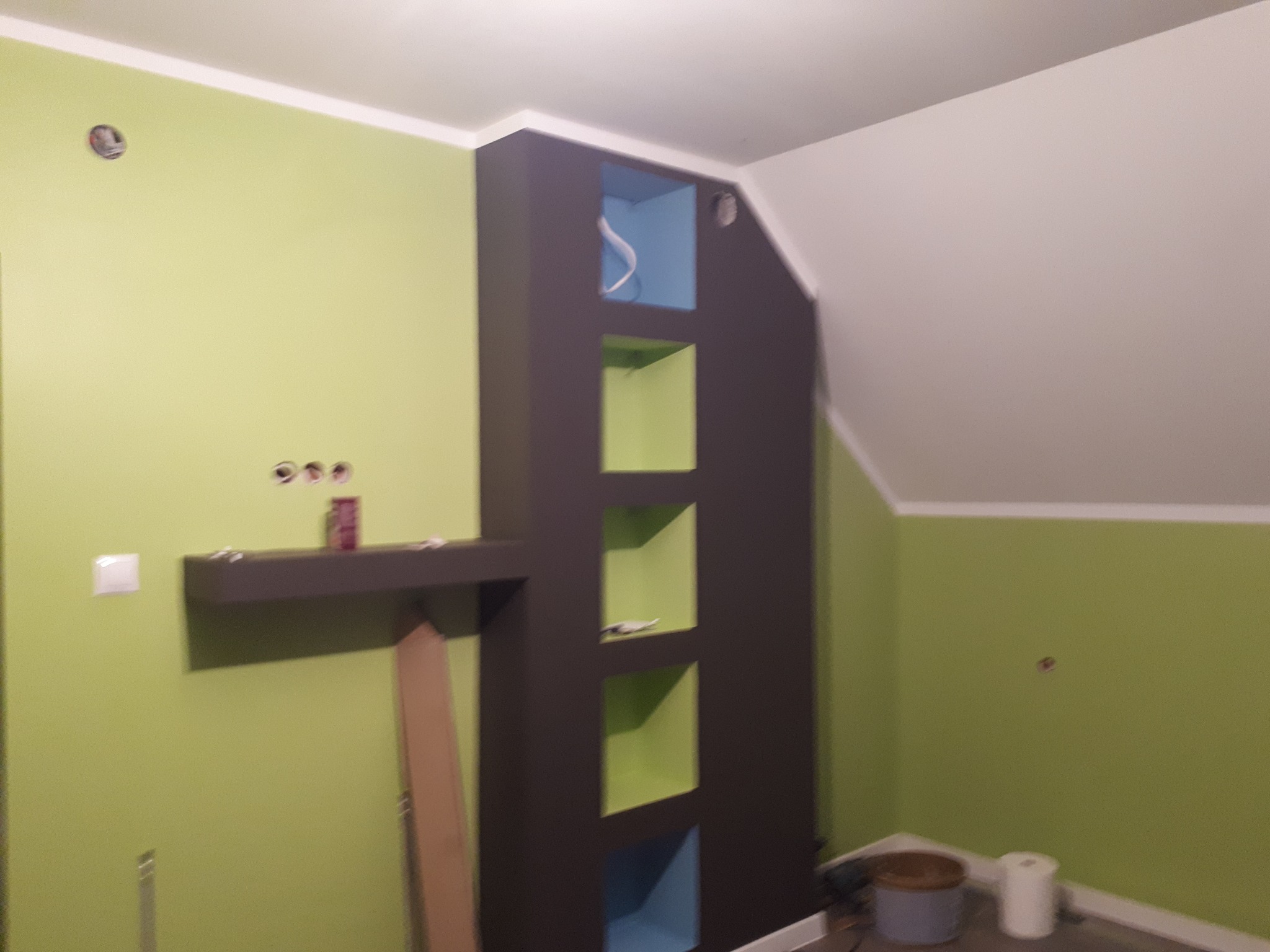 Świeżo pomalowane wnętrze pokoju z wnęką w zabudowie z płyty karton gips w kolorze grafitowym z kolorowymi wstawkami, na ścianie widoczna półka i otwory pod gniazdka.