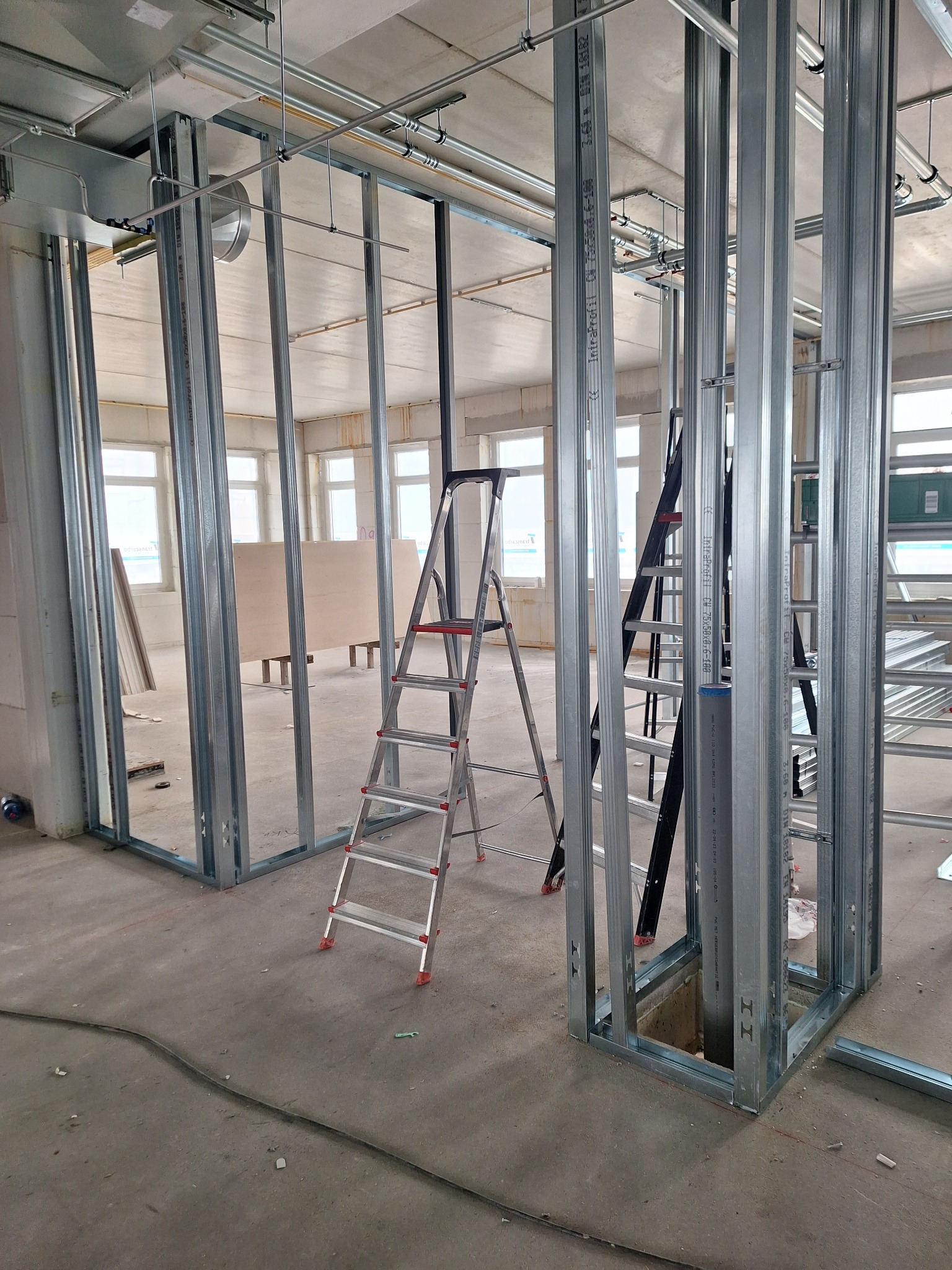 Stelaż z profili stalowych do ścian działowych w surowym stanie, widoczne drabiny, instalacje pod sufitem i okna w tle.