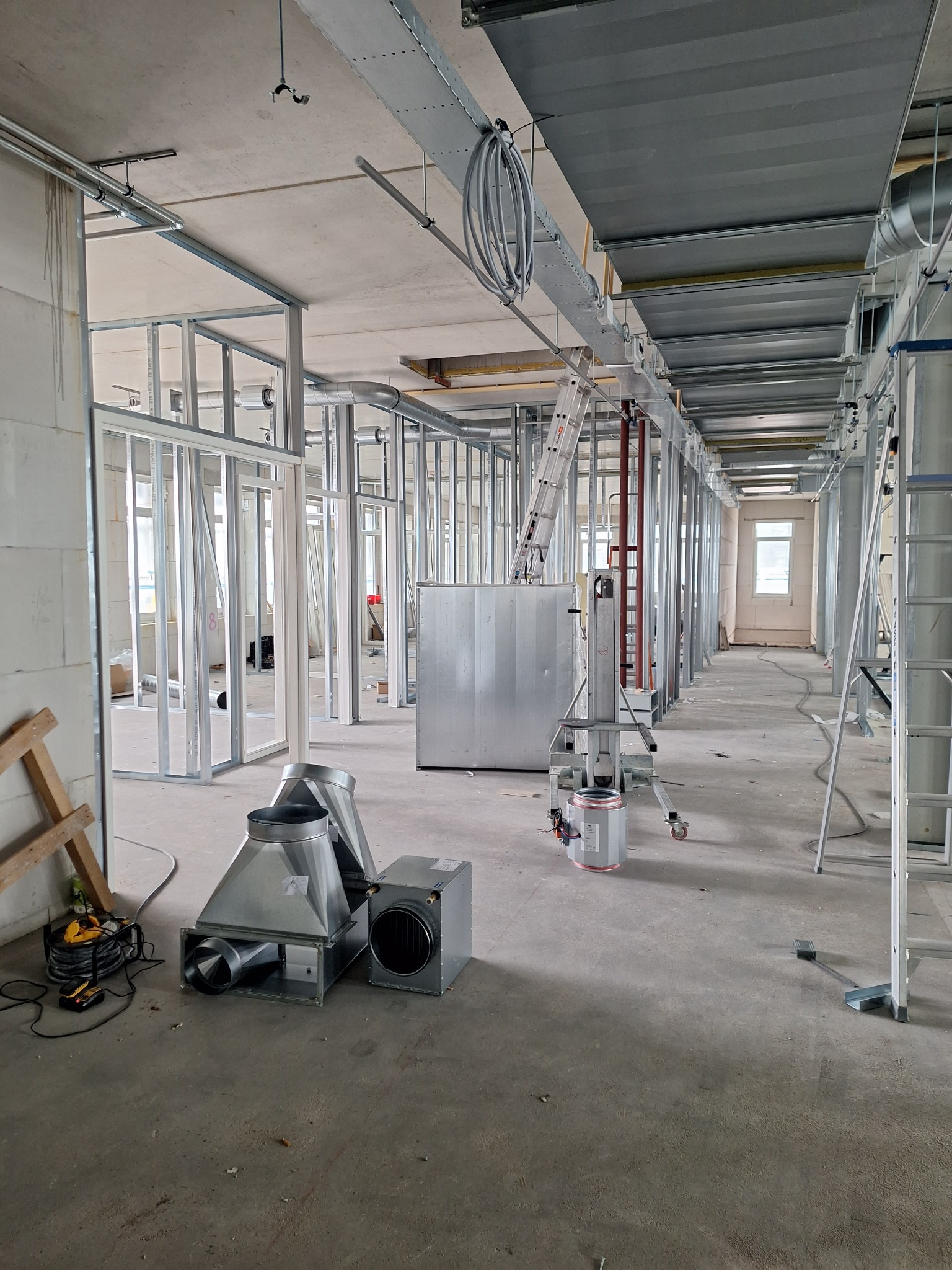 Wnętrze w trakcie prac wykończeniowych: widoczne metalowe profile ścian działowych, elementy wentylacji na podłodze, drabiny i podnośnik, surowy betonowy sufit z instalacjami.