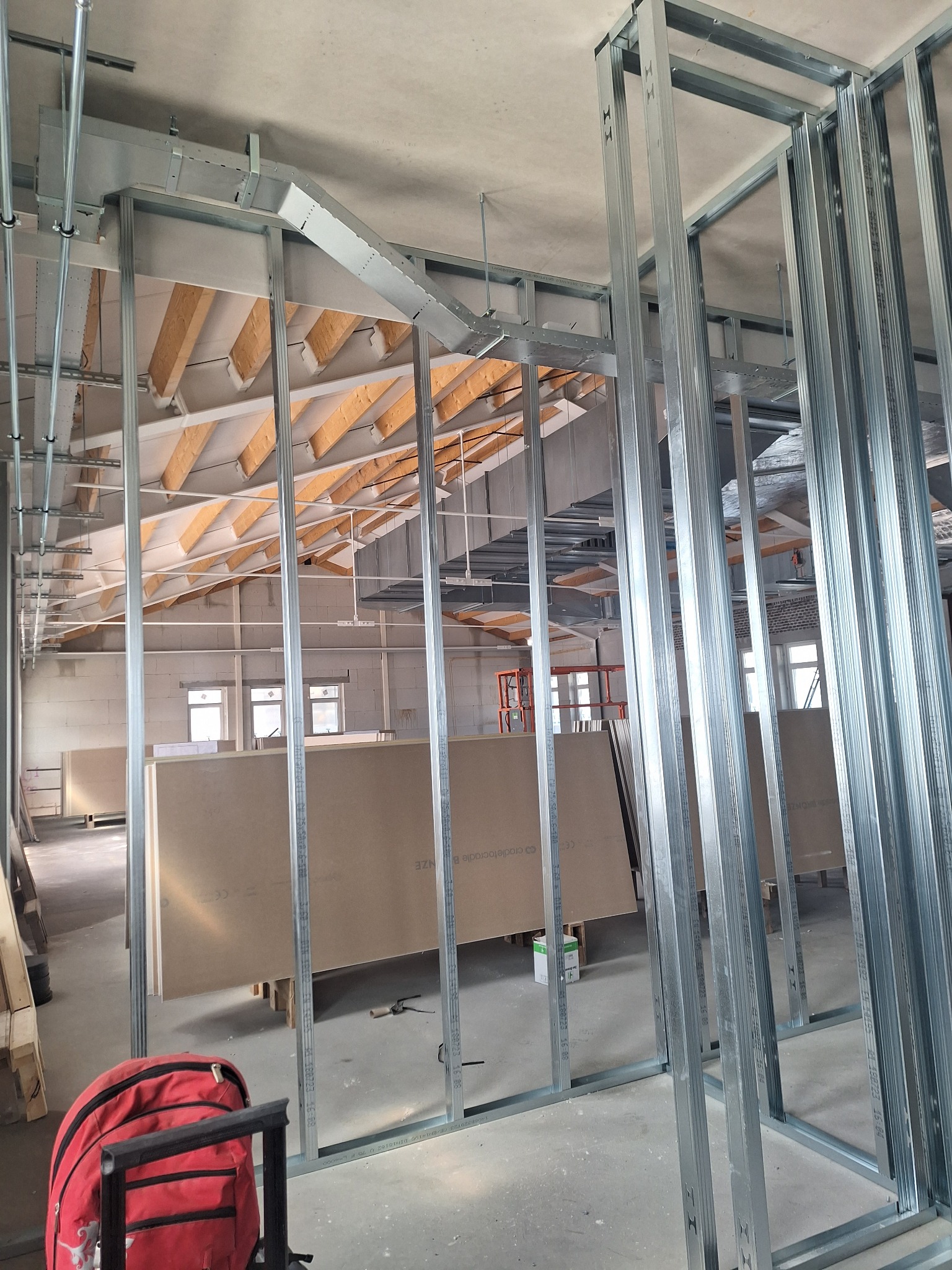 Montaż metalowej konstrukcji szkieletowej ścian działowych w dużym pomieszczeniu z widoczną konstrukcją dachu i płytami gipsowo-kartonowymi, czerwony plecak na pierwszym planie.