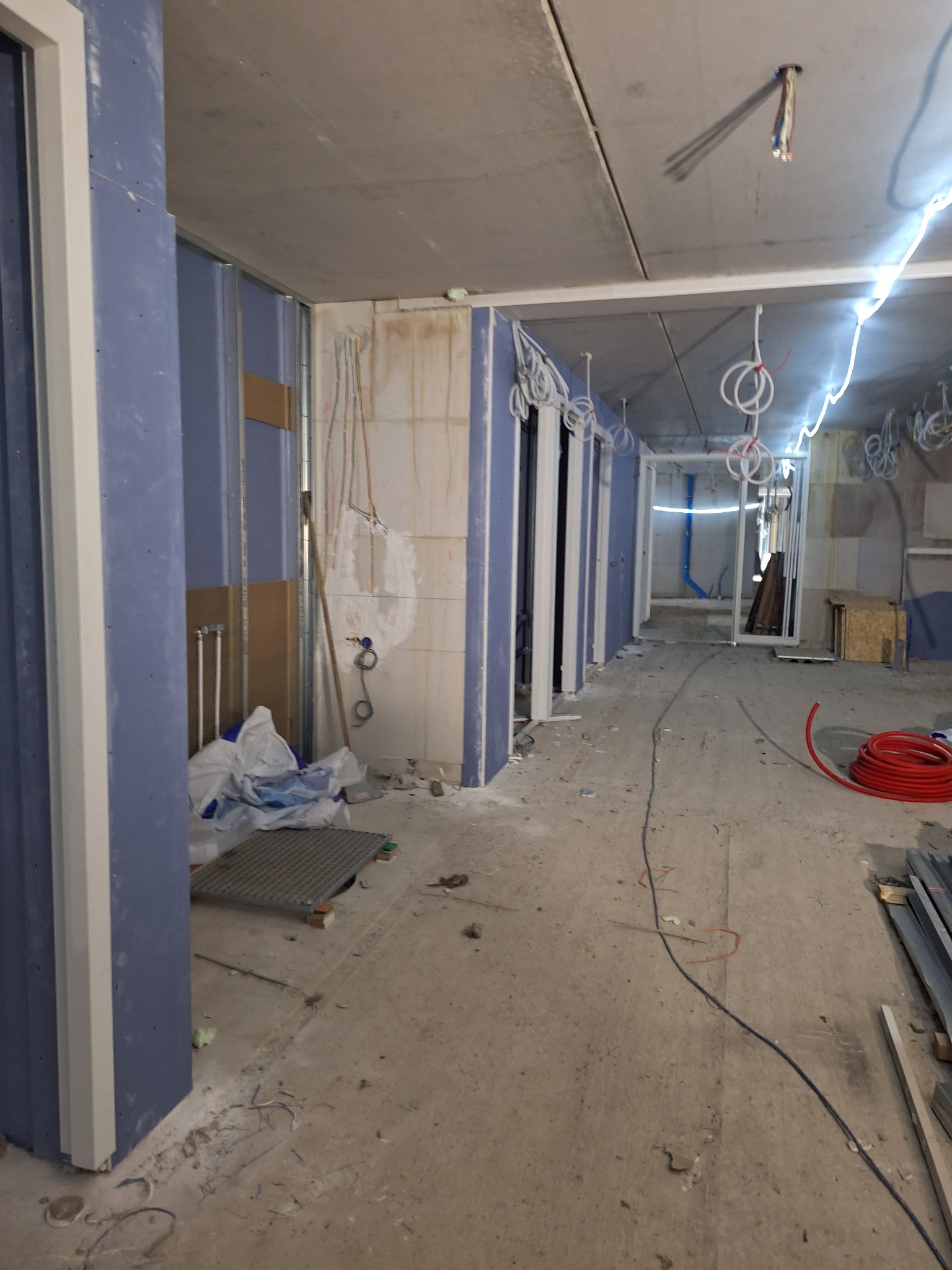 Wnętrze w trakcie prac wykończeniowych: widoczne profile metalowe, płyty karton-gips w kolorze niebieskim, wystające przewody elektryczne i rury, surowy beton na podłodze i suficie, oraz zwisające...