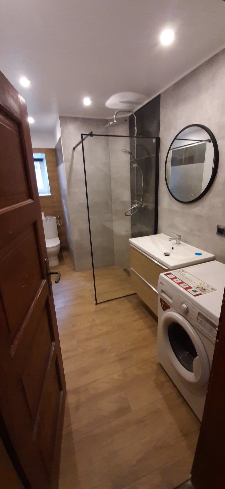 Wykończona łazienka z kabiną prysznicową typu walk-in, okrągłym lustrem, umywalką z szafką i pralką, widok zza otwartych drewnianych drzwi.