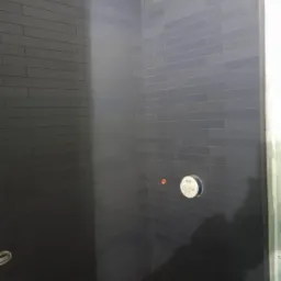 Ciemnoszara, prostokątna glazura ułożona w cegiełkę na ścianach kabiny prysznicowej, widoczny otwór na baterię i przygotowane przyłącze wodne, odpływ liniowy w rogu.