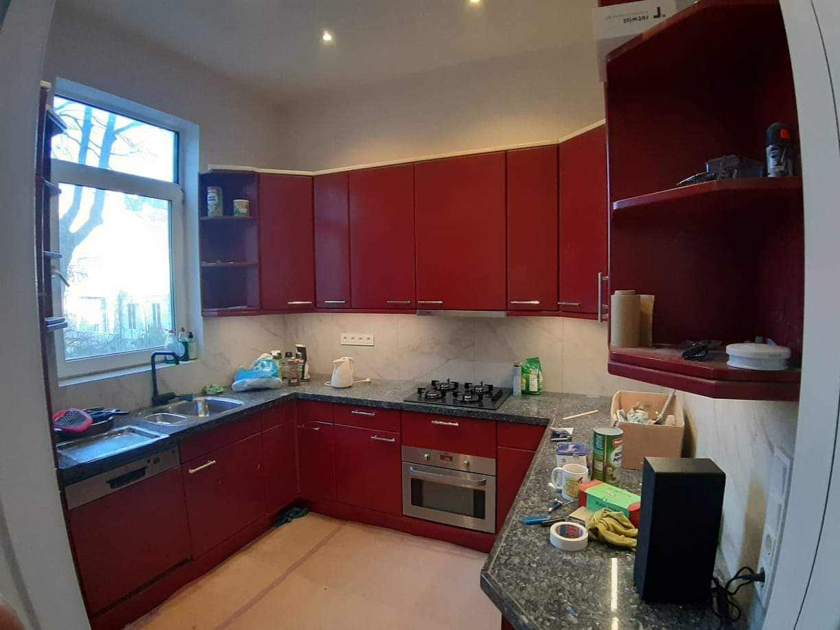 Czerwona kuchnia w trakcie remontu: widoczne narzędzia, niedokończone wykończenie podłogi, okno z widokiem na drzewo, marmurowy blat i częściowo zabudowane szafki.