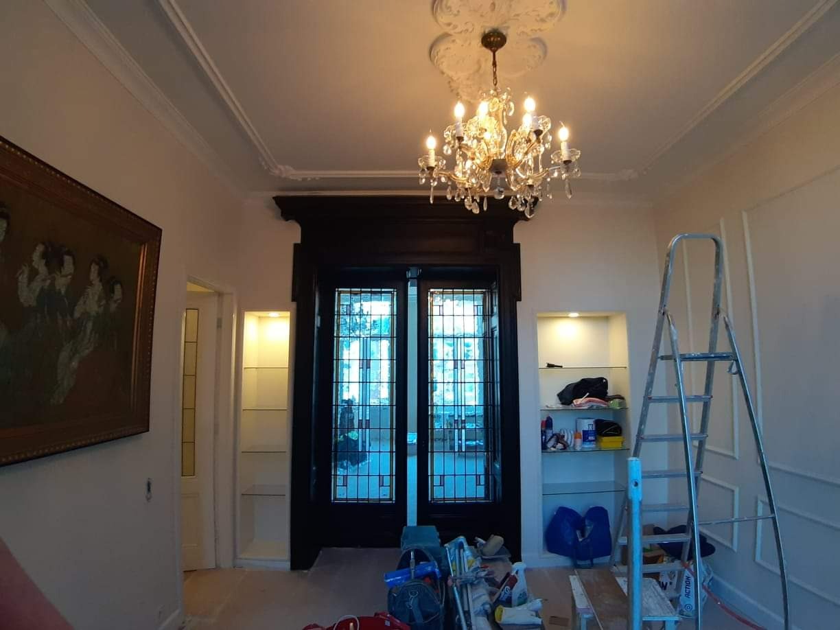 Pokój w trakcie odświeżania, widoczna drabina, podłoga zabezpieczona folią, ściany po malowaniu z dekoracyjnymi listwami, czarne ramy okienne i drzwiowe, kryształowy żyrandol, obraz w złotej ramie.