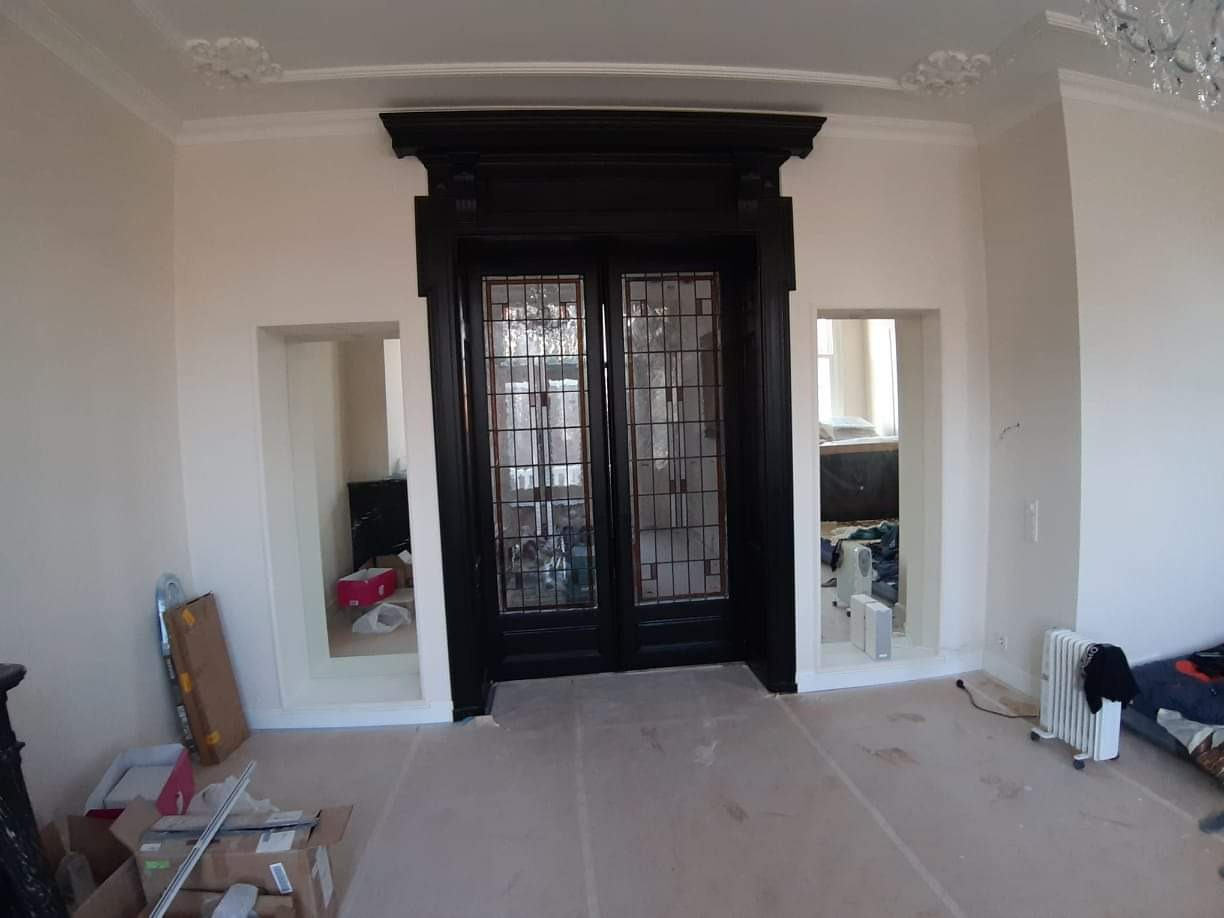 Pokój w trakcie prac wykończeniowych: podłoga zabezpieczona folią, widoczne kartony z materiałami budowlanymi, czarne, dwuskrzydłowe drzwi z szybami w ozdobnej ramie, dekoracyjny sufit z gzymsami.