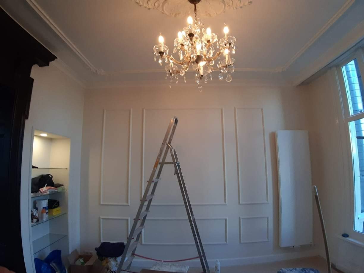 Wnętrze pokoju w trakcie prac wykończeniowych: ściany z dekoracyjnymi listwami, drabina oparta o ścianę, kryształowy żyrandol pod sufitem z dekoracyjnym gzymsem.