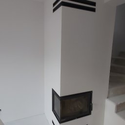 KLAU'S - REMONTY Kamil Szawłowski - Biały narożny kominek z czarnymi akcentami w nowo wykończonym wnętrzu, podłoga zabezpieczona folią malarską, widoczne schody w tle.