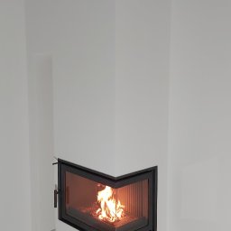 KLAU'S - REMONTY Kamil Szawłowski - Nowoczesny, narożny kominek z czarnym obramowaniem i płomieniami widocznymi przez szybę, w surowym wnętrzu z białymi ścianami i szarą podłogą przed położeniem wykończenia.