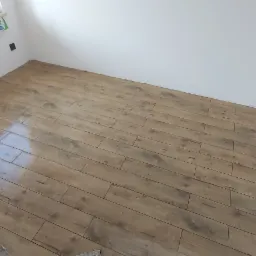 Ukończona podłoga z paneli imitujących drewno w jasnym pomieszczeniu z białymi ścianami; widoczny fragment ściany w stanie surowym.