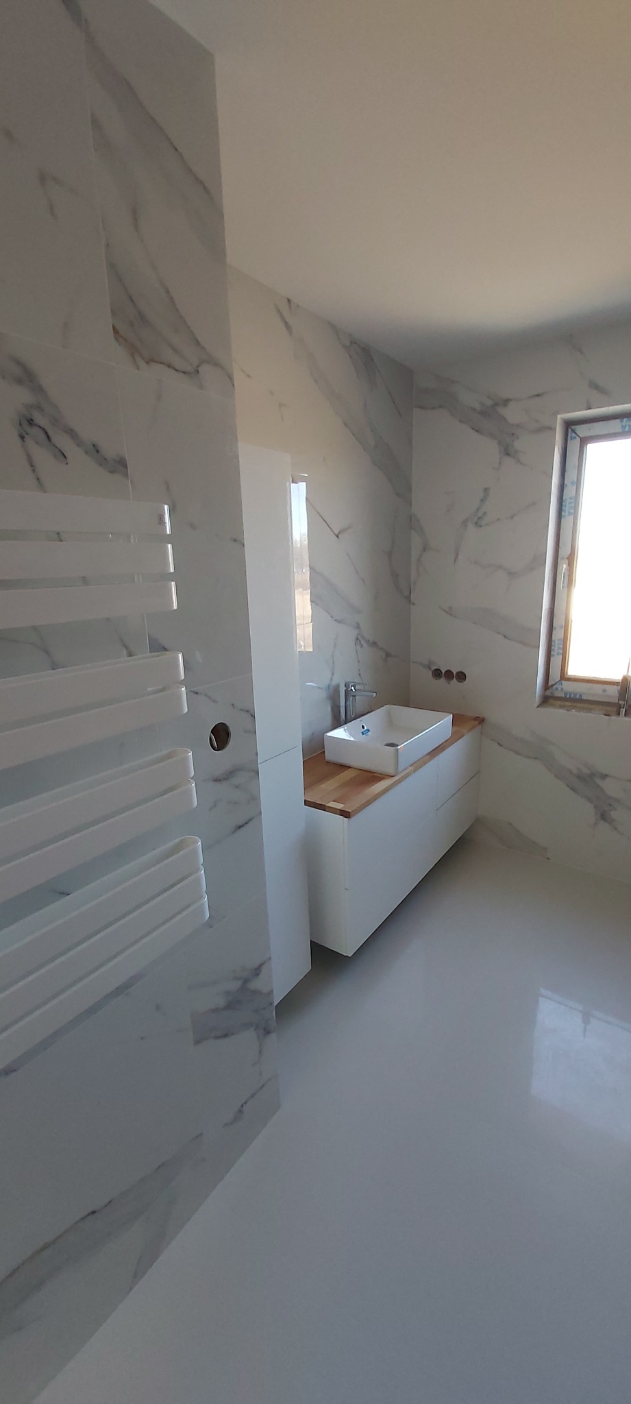 Biała łazienka z marmurowymi płytkami na ścianach, grzejnikiem drabinkowym i umywalką nablatową na drewnianym blacie.