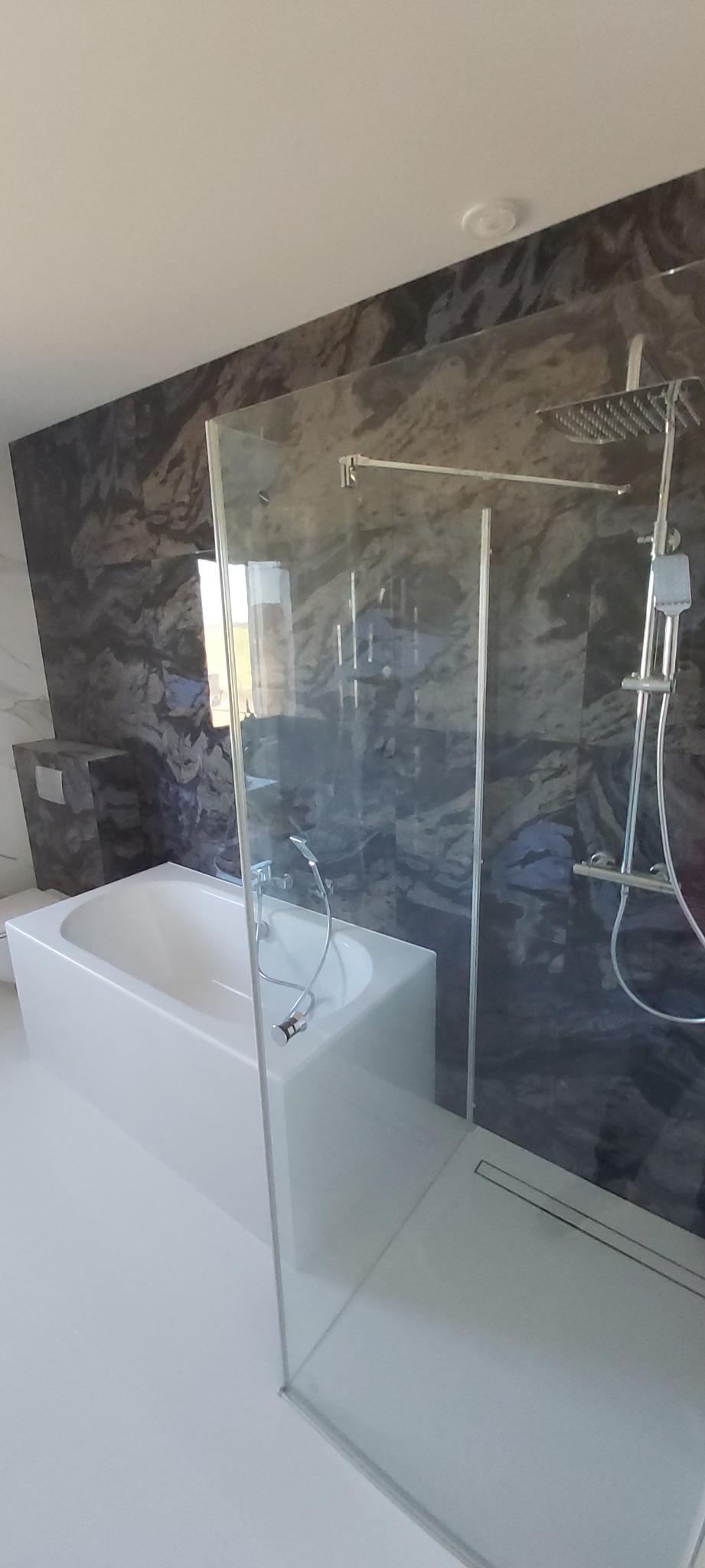 Nowoczesna łazienka z kabiną prysznicową typu walk-in, białą wanną i ścianą wykończoną płytkami imitującymi marmur w odcieniach szarości i granatu. Widoczny odpływ liniowy w posadzce.