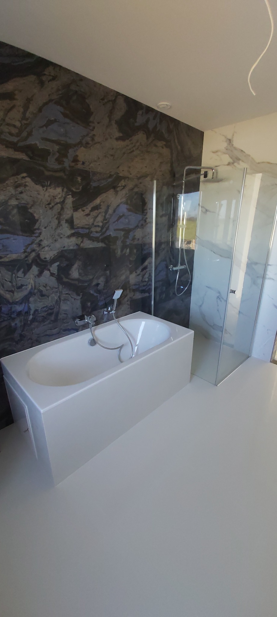 Nowoczesna łazienka z białą wanną, szklanym prysznicem i ścianami wyłożonymi płytkami imitującymi marmur w odcieniach szarości i granatu.