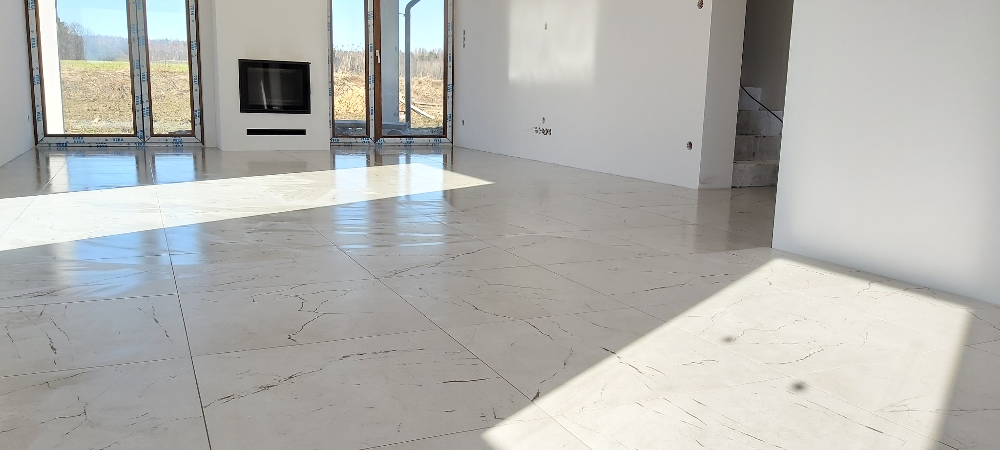 Jasne, przestronne wnętrze z nową, błyszczącą podłogą z płytek imitujących marmur, w tle widoczne okna z widokiem na zewnątrz i surowy kominek.