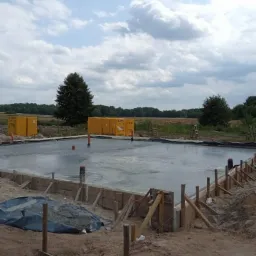 Wylany fundament pod budowę domu, widoczne szalunki i wystające rury, w tle palety z materiałami budowlanymi i krajobraz polny pod pochmurnym niebem.