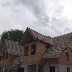 Wznoszenie dwukondygnacyjnego domu z czerwonej cegły ceramicznej, widoczne krokwie dachu pokryte folią, drabina oparta o ścianę, pochmurne niebo w tle.