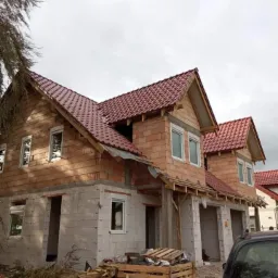 Budowa domu dwukondygnacyjnego w stanie surowym otwartym, widoczne ściany z bloczków i cegły, czerwona dachówka, okna wstawione, teren wokół budowy zagracony drewnem i odpadami, częściowo widoczny...