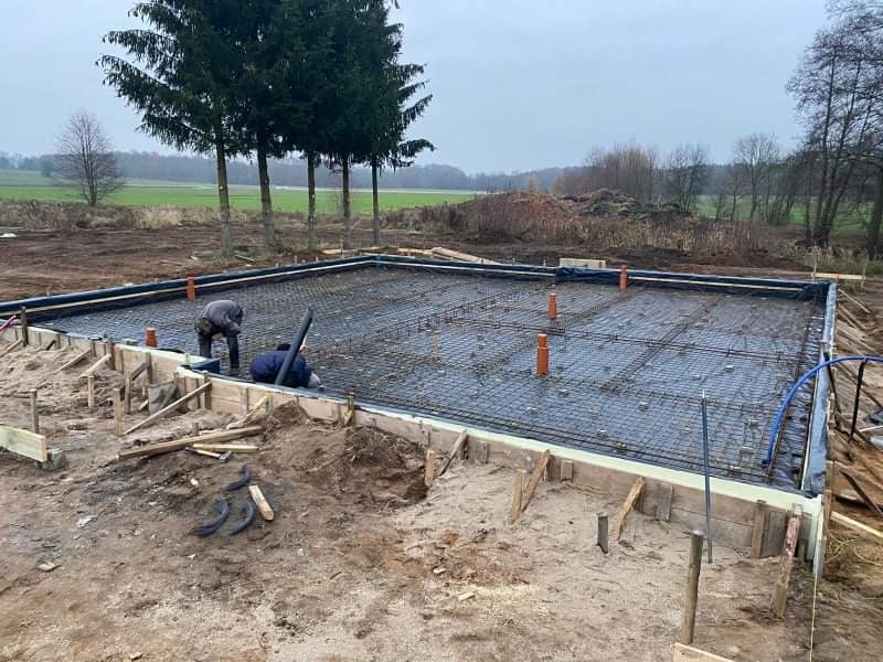 Przygotowanie fundamentów pod budowę domu: zbrojenie, szalunek drewniany i izolacja przeciwwilgociowa, widoczni dwaj pracownicy w trakcie montażu rur kanalizacyjnych.