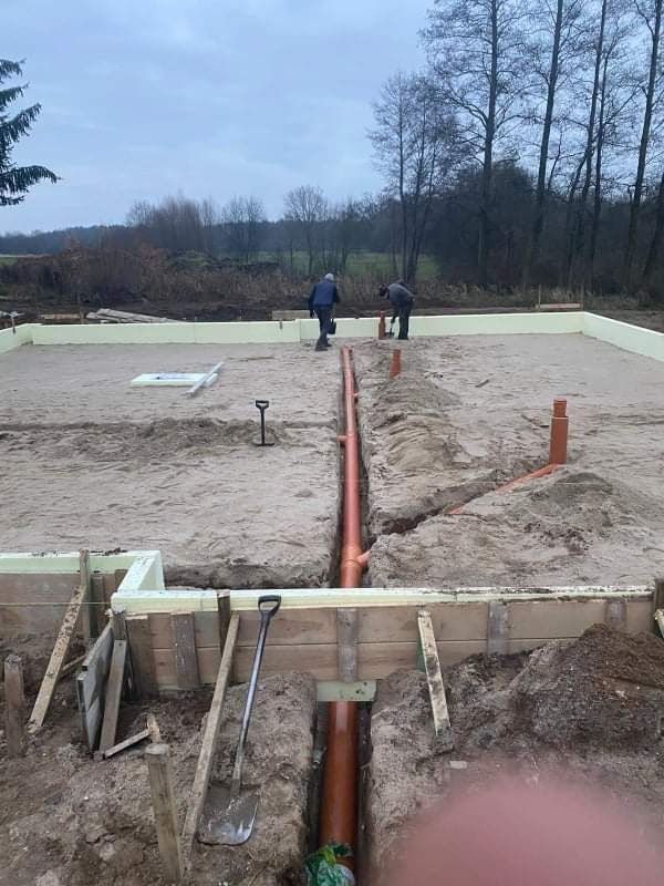 Fundamenty domu w trakcie budowy z widoczną izolacją termiczną i ułożonymi rurami kanalizacyjnymi w wykopach, dwóch pracowników w tle.