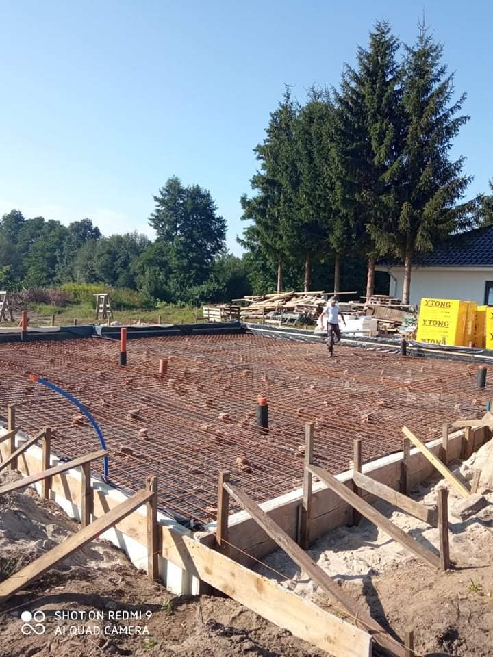 Zbrojenie fundamentów domu jednorodzinnego z widocznymi szalunkami, rurami instalacyjnymi i paletami bloczków Ytong w tle.