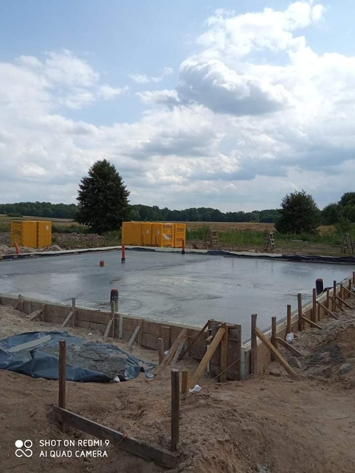 Wylany fundament pod budowę domu, widoczne szalunki i wystające rury, w tle palety z materiałami budowlanymi i krajobraz polny pod pochmurnym niebem.