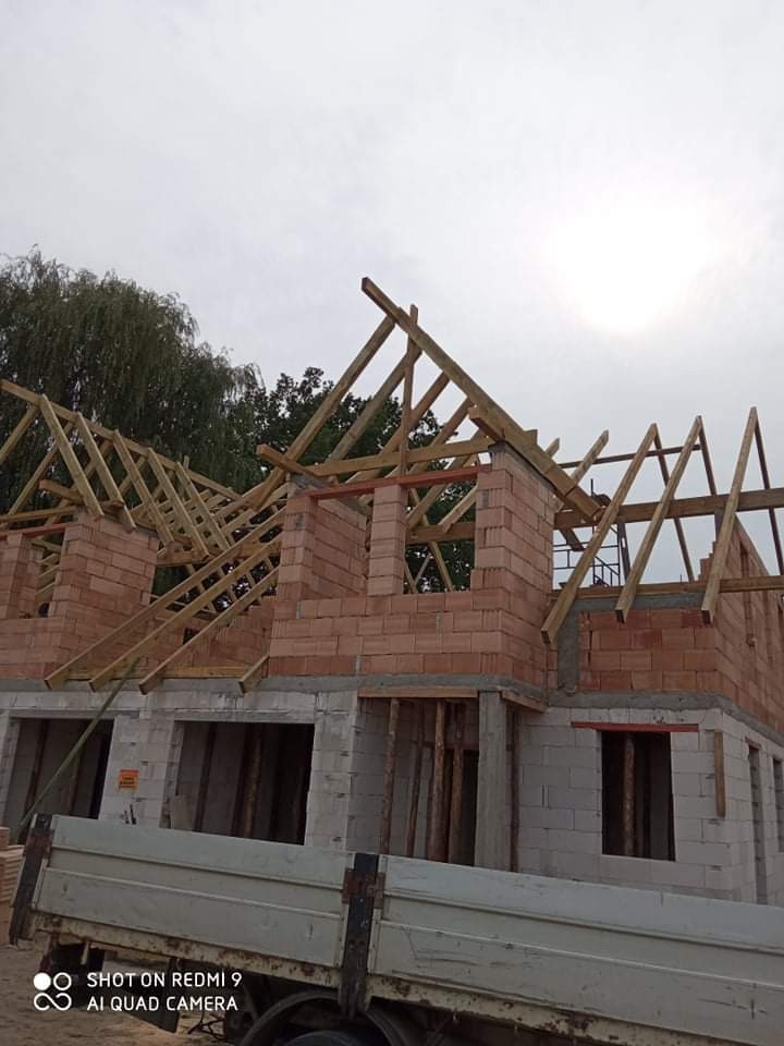 Wznoszenie ścian z czerwonej cegły i konstrukcji drewnianej dachu na budowie domu jednorodzinnego, widok z perspektywy skrzyni ładunkowej samochodu dostawczego.