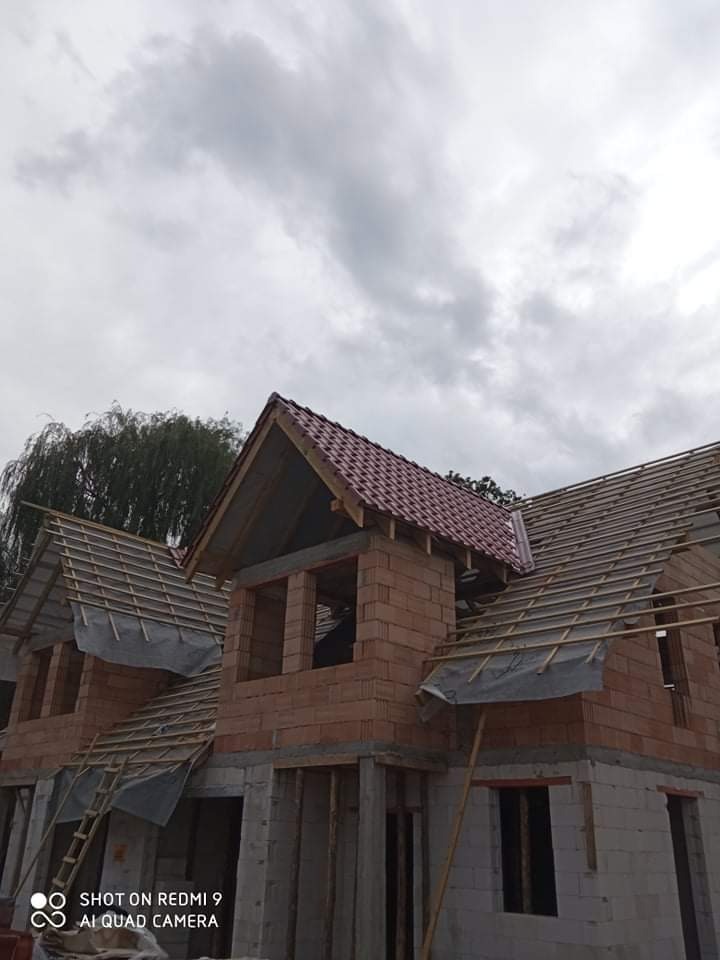Wznoszenie dwukondygnacyjnego domu z czerwonej cegły ceramicznej, widoczne krokwie dachu pokryte folią, drabina oparta o ścianę, pochmurne niebo w tle.