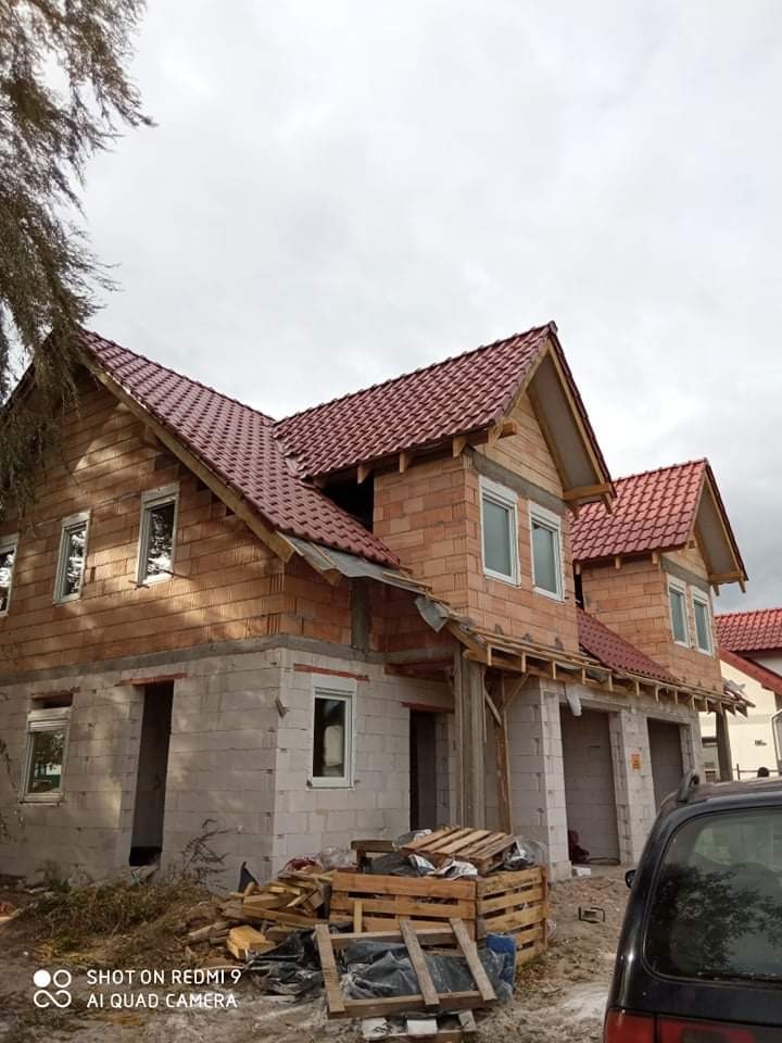 Budowa domu dwukondygnacyjnego w stanie surowym otwartym, widoczne ściany z bloczków i cegły, czerwona dachówka, okna wstawione, teren wokół budowy zagracony drewnem i odpadami, częściowo widoczny...