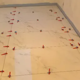 Układanie płytek imitujących marmur na podłodze z użyciem systemu poziomowania z czerwonymi klinami i niebieskimi nakładkami.