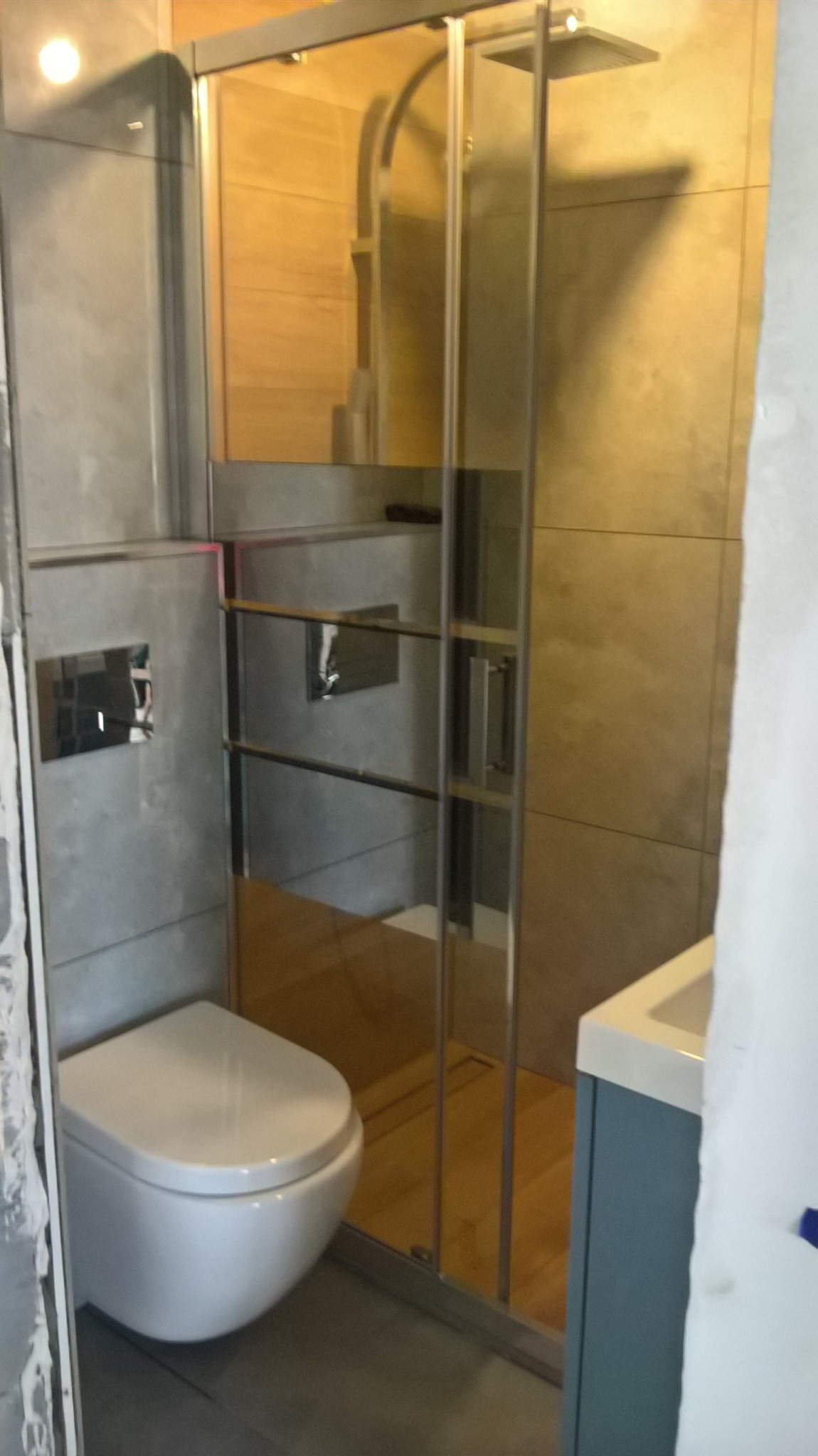 Nowoczesna łazienka z podwieszaną toaletą, szklaną kabiną prysznicową walk-in i umywalką na szafce w odcieniach szarości i drewna.