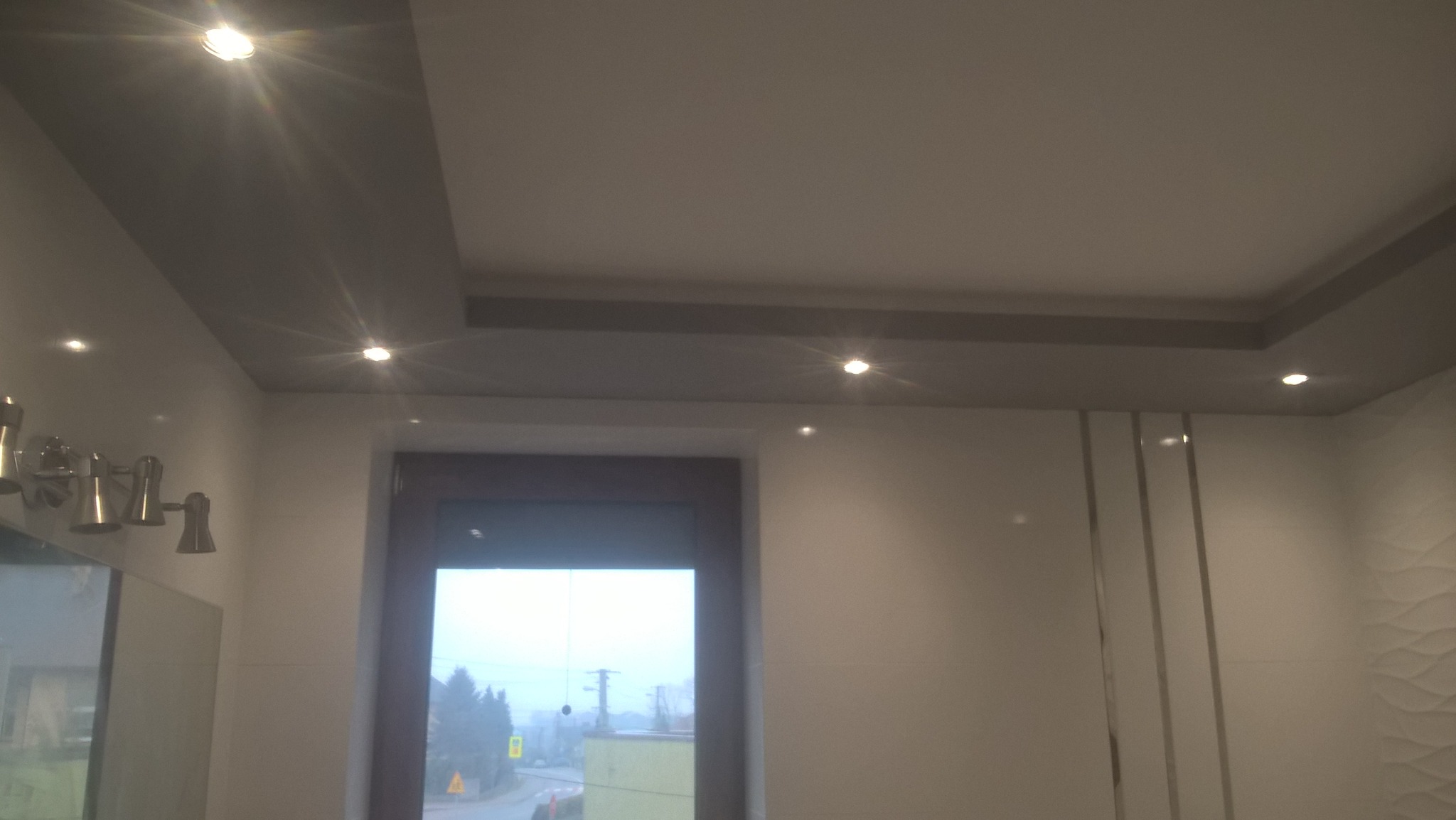 Wykończone wnętrze łazienki z podwieszanym sufitem z oświetleniem punktowym, widok na okno i dekoracyjną ścianę z subtelnym wzorem fal.