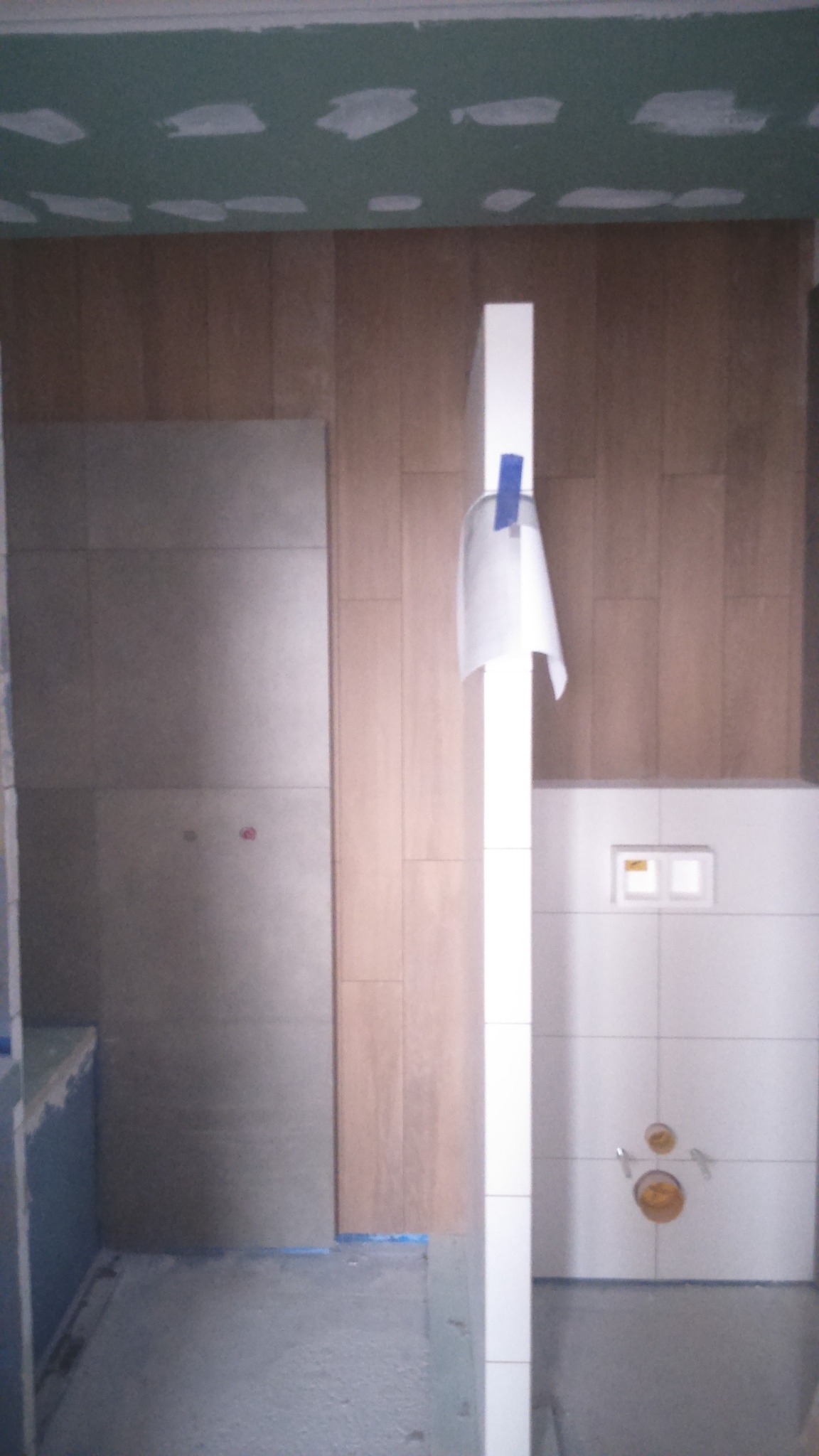 Niedokończona łazienka z częściowo ułożonymi płytkami imitującymi drewno na ścianie, surowy sufit z widocznymi łatami gipsowymi, białe płytki na ścianie z zamontowaną armaturą i białą kolumną...