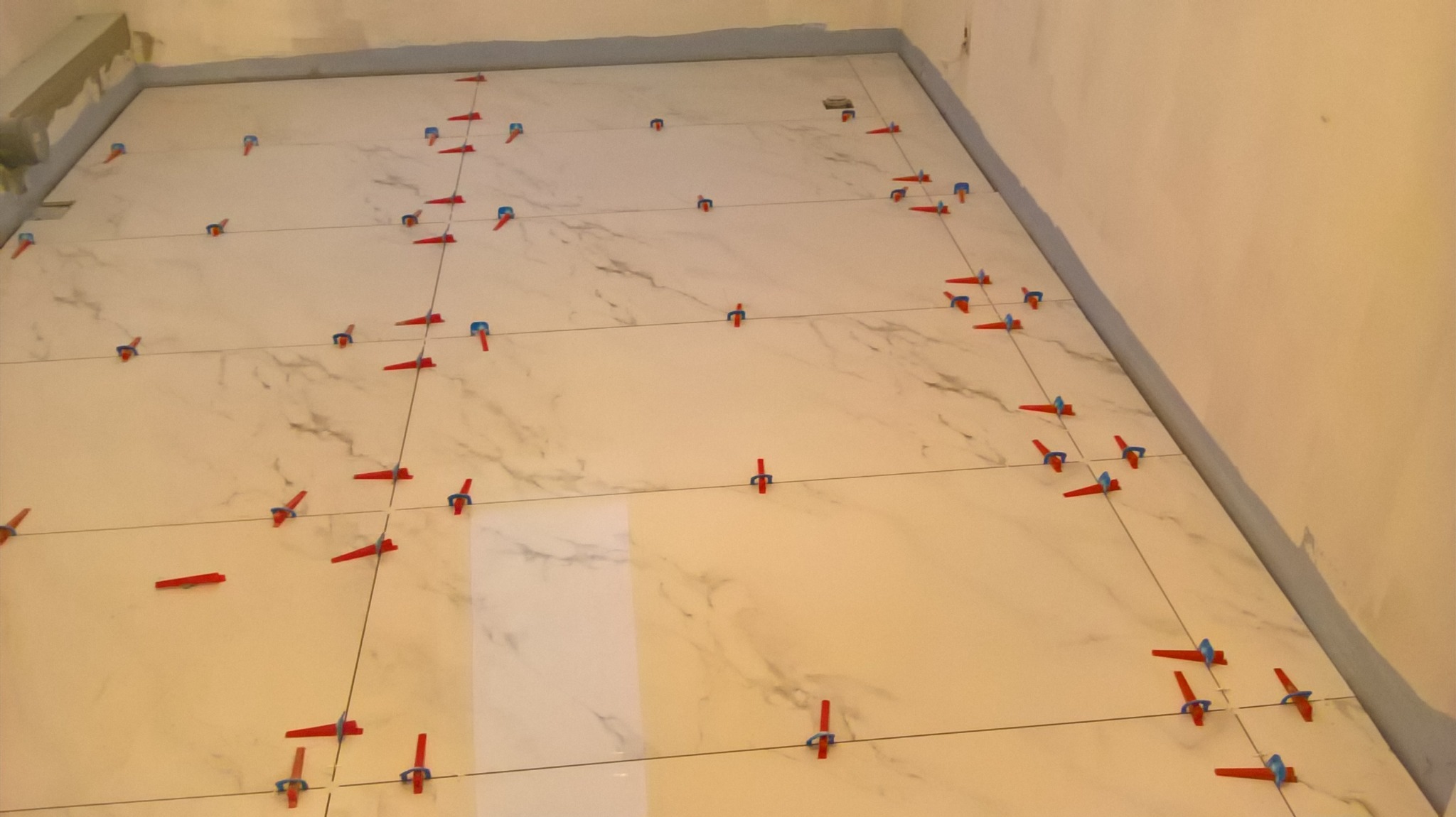 Układanie płytek imitujących marmur na podłodze z użyciem systemu poziomowania z czerwonymi klinami i niebieskimi nakładkami.