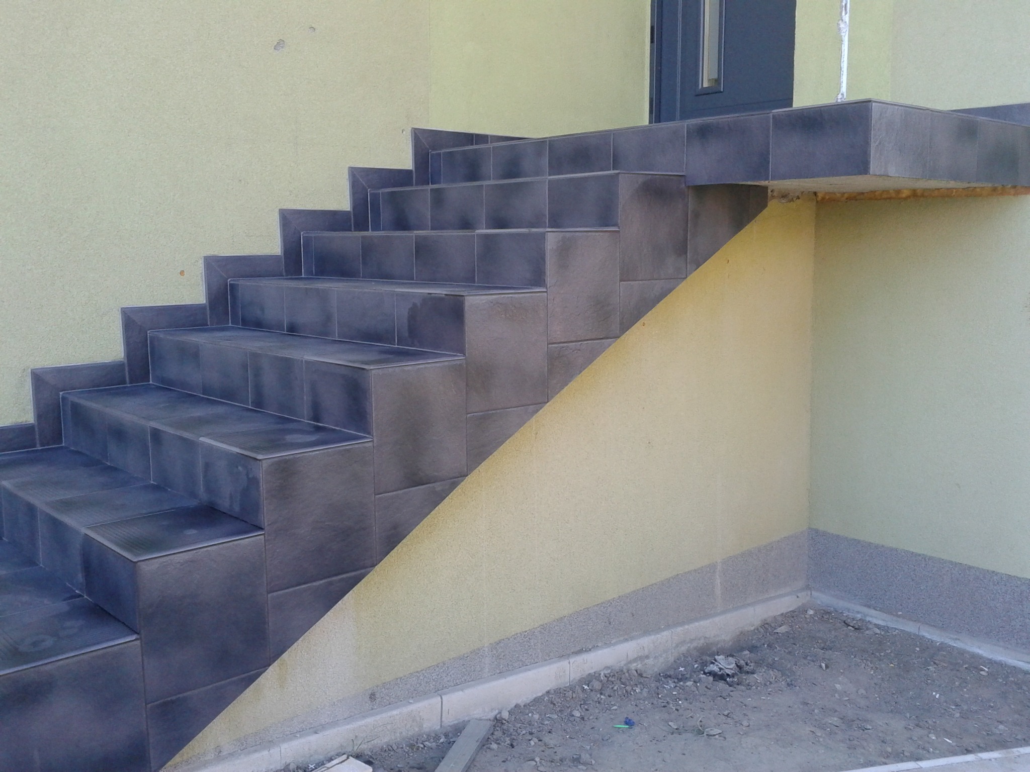 Zewnętrzne schody wejściowe wyłożone ciemnymi, kwadratowymi płytkami, z widocznymi fugami i fragmentem elewacji budynku w kolorze żółtym.