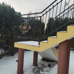 Zewnętrzne schody z żółtymi stopniami i balustradą z czarnego metalu z dekoracyjnymi, złotymi elementami na tle zimowego ogrodu.